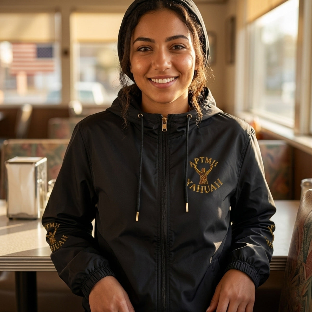 A smiling woman in a black windbreaker featuring a gold logo and "YAHUAH" text is pictured sitting in a sunlit American diner, based on the provided photo.