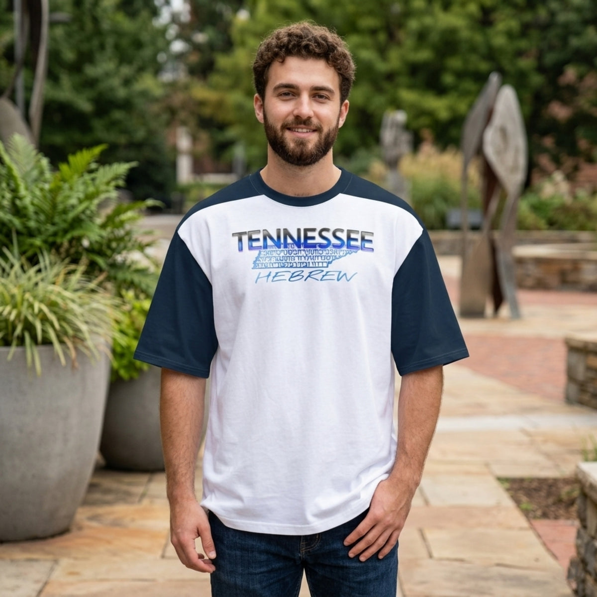 A man wearing a white and navy raglan shirt with a "TENNESSEE HEBREW" graphic featuring a state silhouette on the chest, standing in an outdoor plaza.