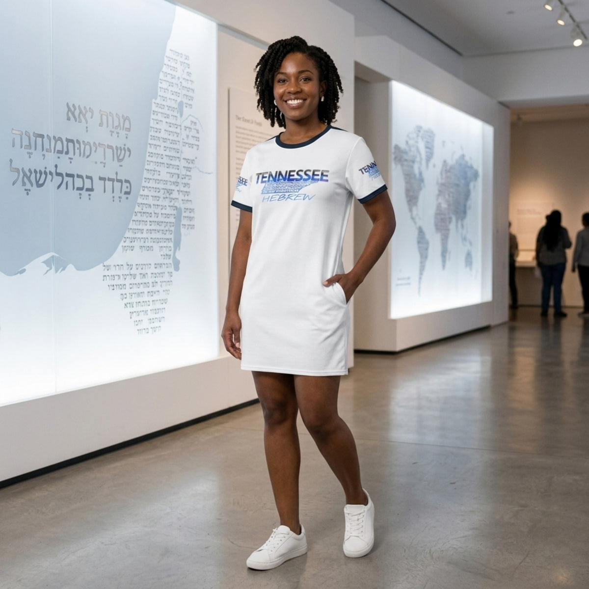 A woman in a white "Tennessee Hebrew" t-shirt dress smiles in a museum with Hebrew text displays and a world map in the background.